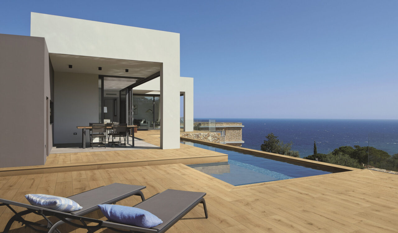 Wood-look natural stone deck with lounge chairs beside an infinity pool overlooking the ocean at a modern white villa.