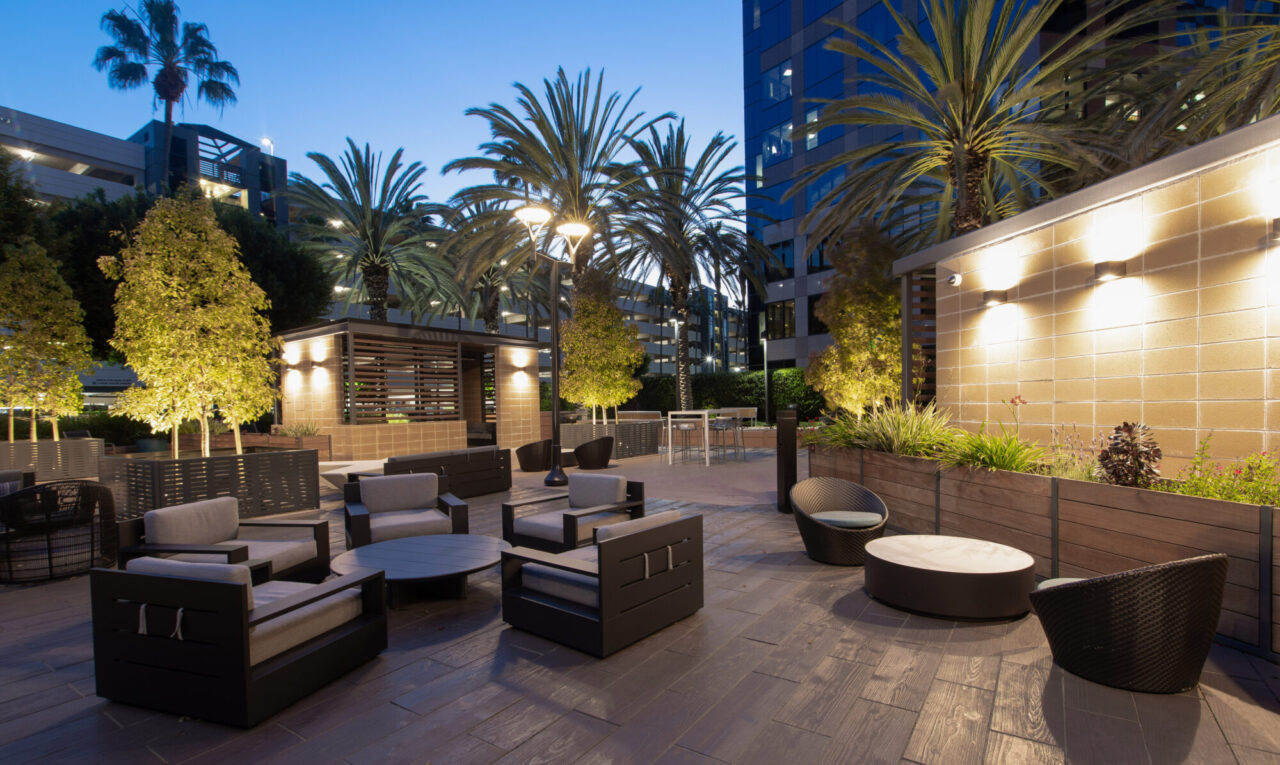 Outdoor lounge cabana with concrete pillars, slatted wood pergola roof, wall-mounted TV, and dark modern sofas, illuminated by sconce lighting, set on a striped paver plaza with planter boxes and palm trees at dusk.