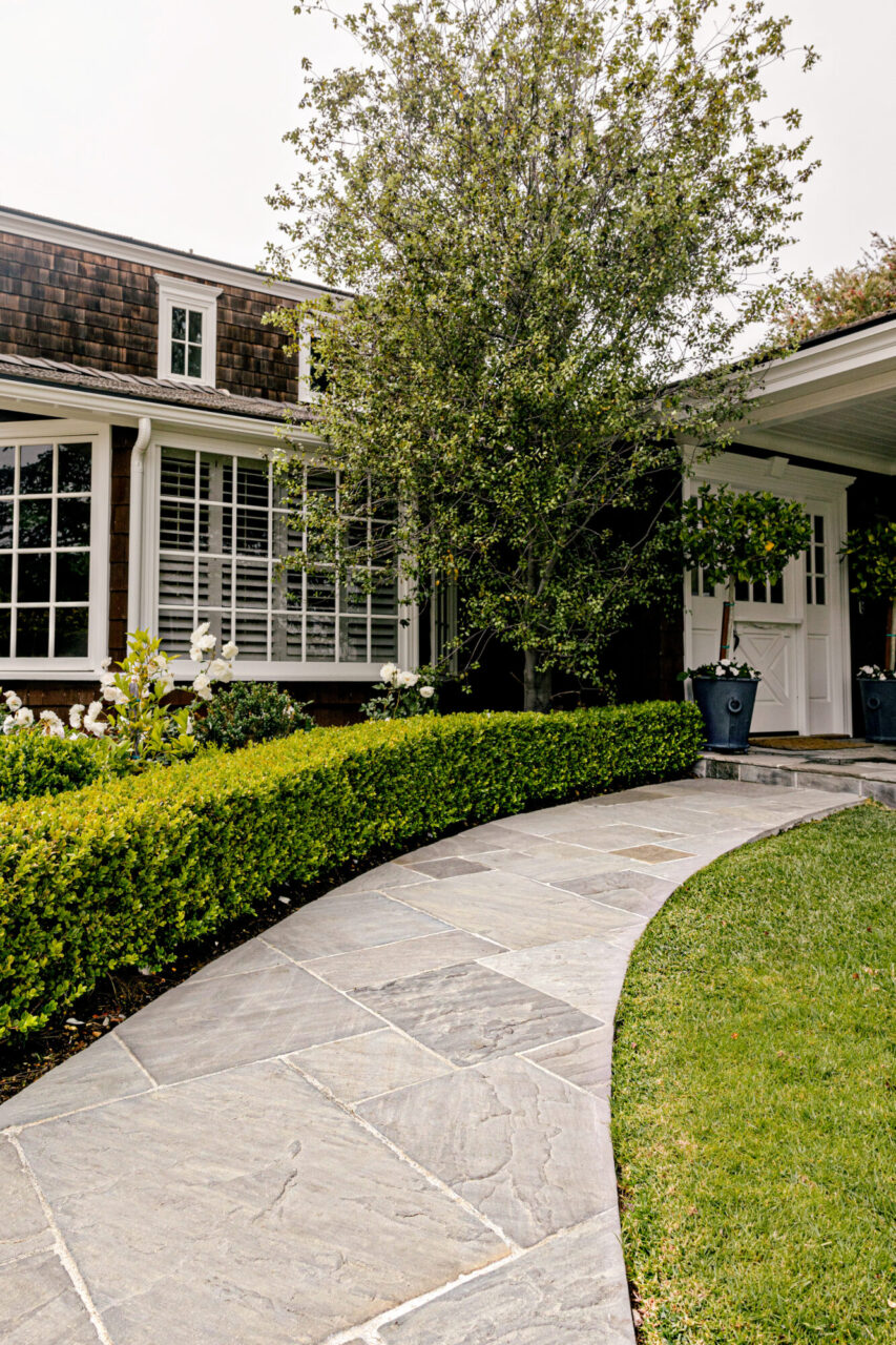 Curved natural stone paver walkway lined with a manicured hedge leading to a shingle-style home with a white barn door entrance.
