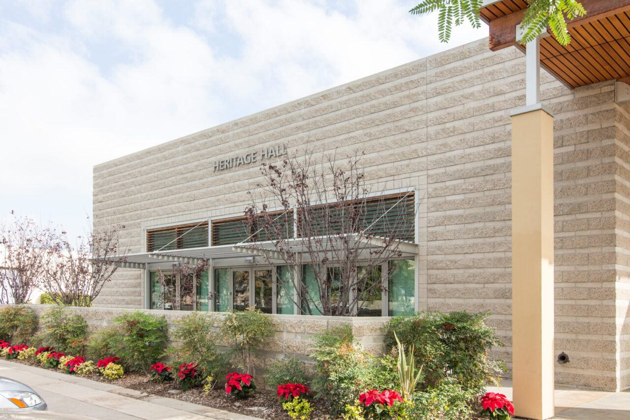 Modern building entrance with a wood-slat overhang supported by tall square columns, glass doors, mixed concrete block and wood siding, shaded by a large overhanging tree.