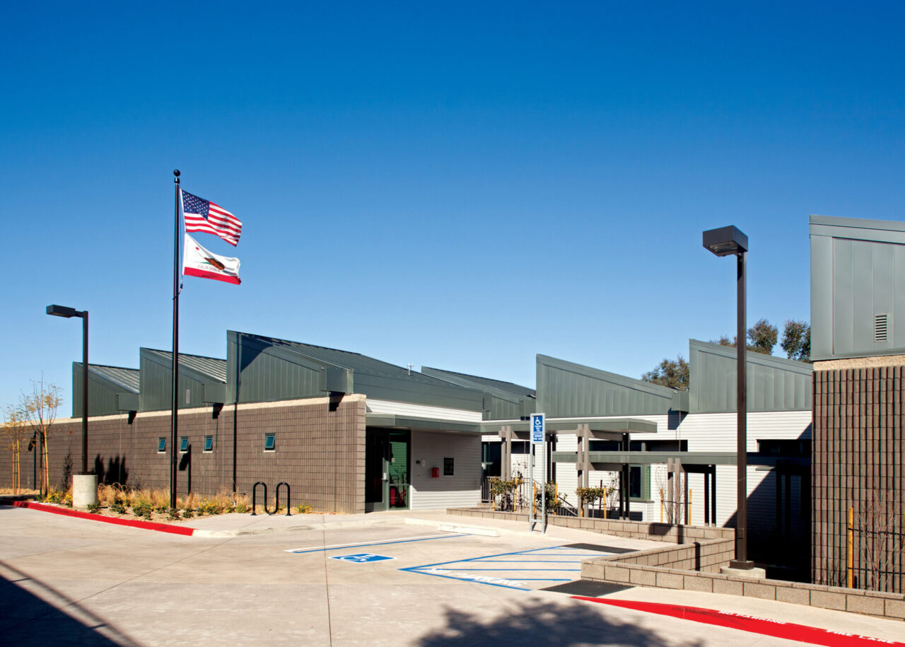 Public building exterior with a saw-tooth metal roof, scored concrete block walls, American and California flags on a pole, accessible parking spaces, and bike racks in the foreground under a clear blue sky.