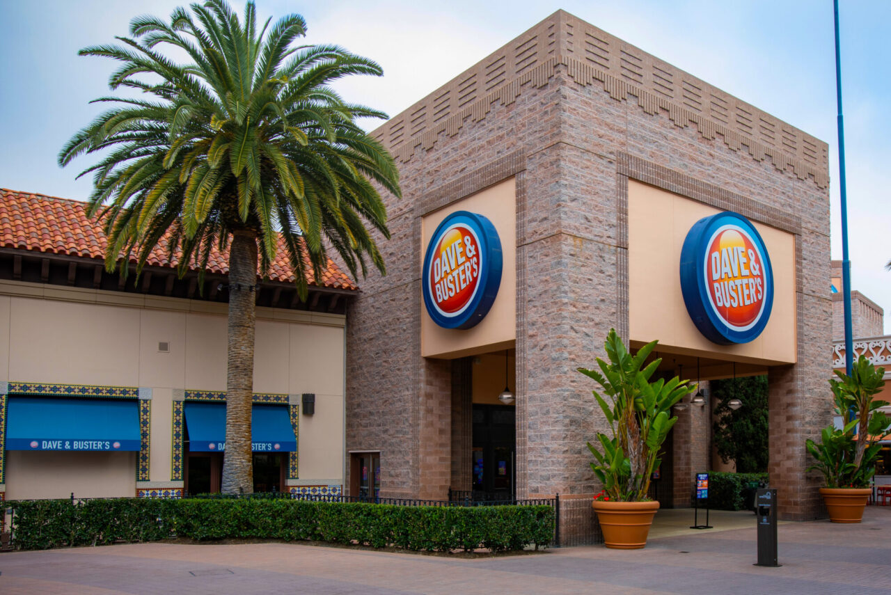 Corner concrete block exterior of a Dave & Buster's building with two large circular logo signs, flanked by a tall palm tree, potted plants, and a Spanish-tile-roofed storefront with blue awnings in a shopping plaza.