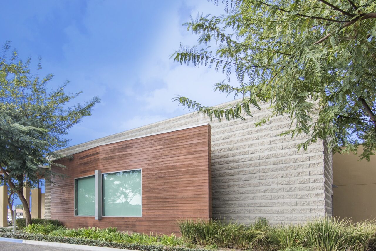 Modern building exterior with a curved roofline transitioning between reddish wood-slat siding and light textured concrete block, a large window, ornamental grasses along the foundation, and overhanging tree branches.