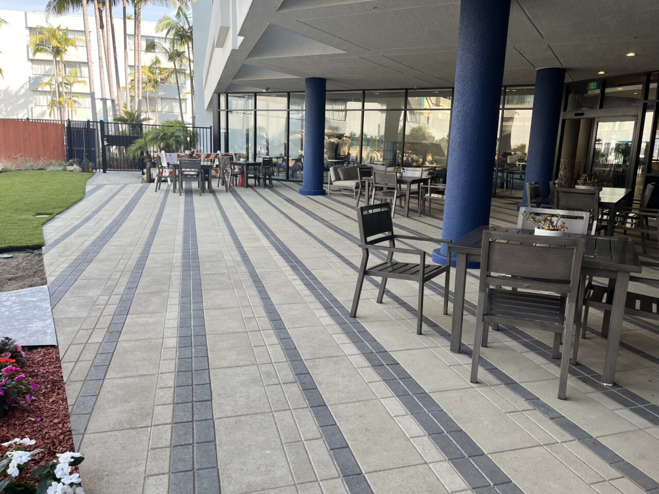Hotel outdoor patio with striped gray paver flooring, gray metal dining chairs and tables under a covered walkway with blue columns, a glass building facade, palm trees, and a flowering garden bed in the foreground.