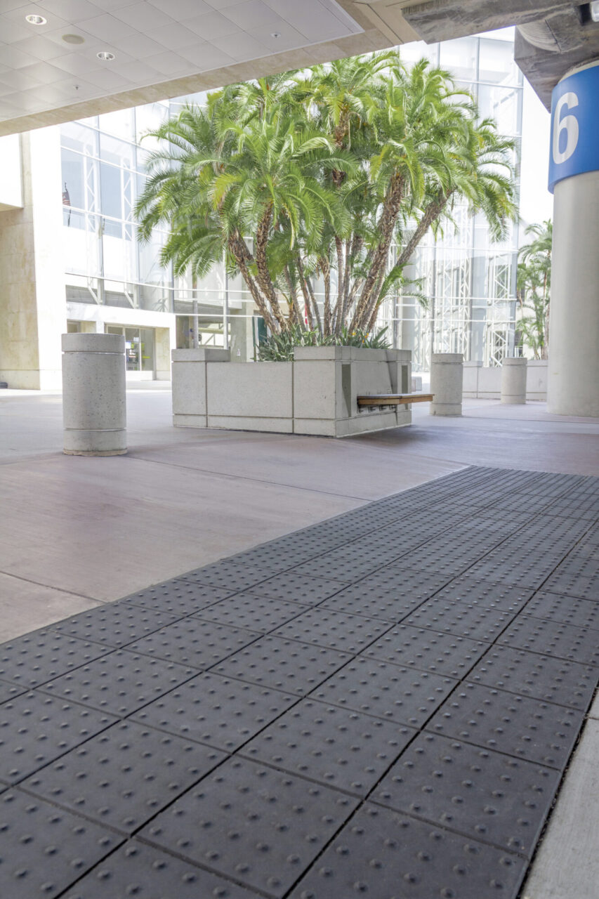 Airport terminal covered walkway with textured tactile paving, concrete bollards, and a raised planter box with tall palm trees and an integrated wood bench, in front of a glass building facade.