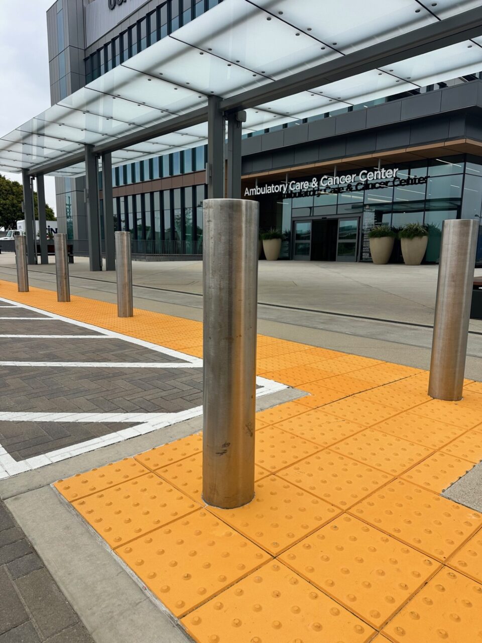 Ambulatory Care and Cancer Center building entrance with a glass canopy walkway, stainless steel bollards, yellow tactile paving, and large potted plants flanking the glass double doors.