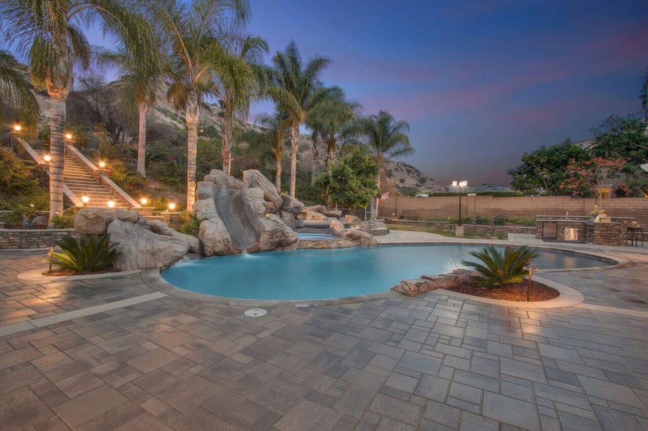 Freeform pool with a boulder water slide and lit staircase at dusk, surrounded by a paver patio and hillside palm trees.