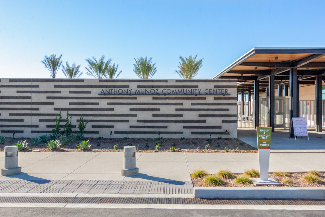 Anthony Munoz Community Center entrance with a striped stone and dark tile monument wall sign, drought-tolerant landscaping with succulents and grasses, bollard lighting, and a covered pick-up drop-off area beneath a modern wood-and-steel canopy.