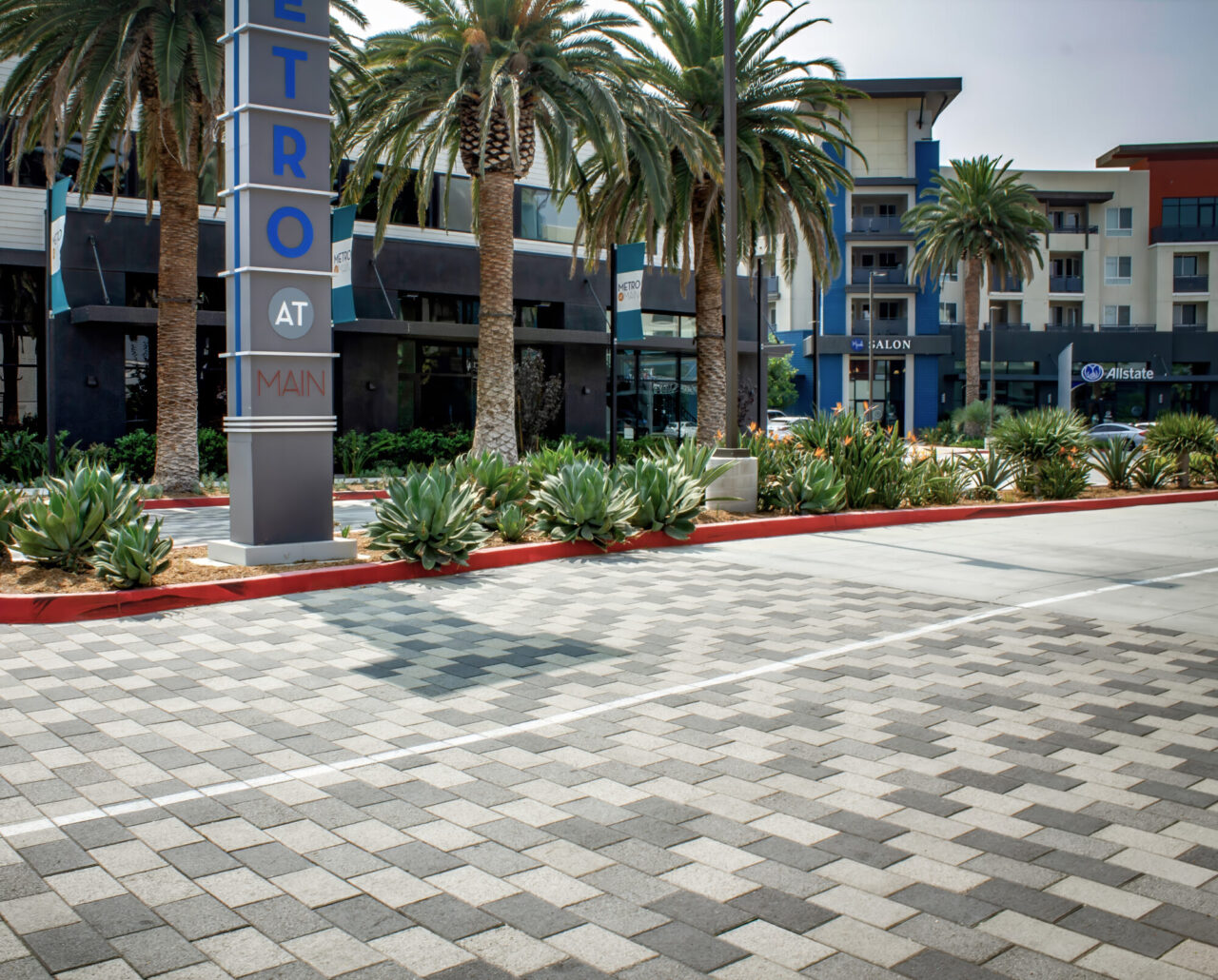 Mixed-use "Metro at Main" development with a tall pylon sign, palm trees, agave landscaping, and storefronts including a salon and Allstate office, set behind a paver street with painted lane markings.