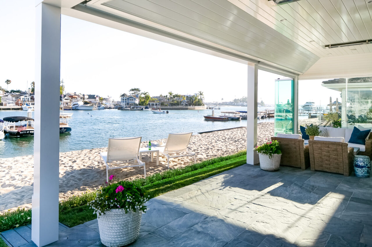 Waterfront patio with lounge chairs on a private sandy beach, overlooking a harbor filled with boats and waterfront homes.