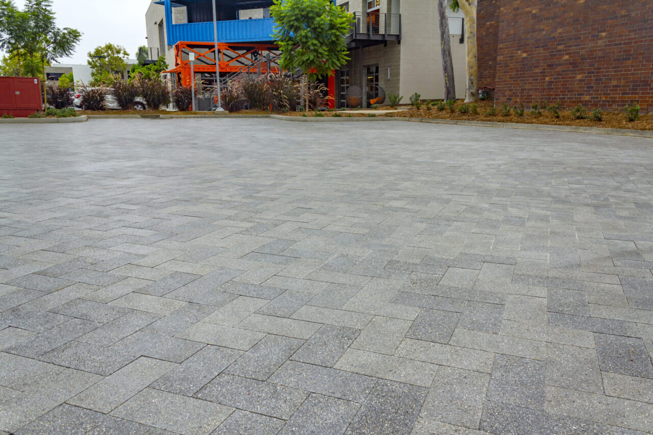 Herringbone-patterned gray paver plaza in front of a mixed-material building with brick and blue metal siding, an orange metal staircase accent, ornamental grasses, and a red mulch planting bed along the building's edge.