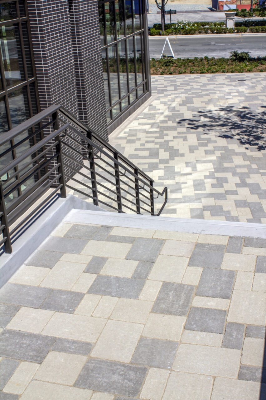 Elevated view of a paved plaza with a random-pattern gray and cream stone tile design, black metal handrails alongside stairs, and a brick-and-glass building facade in the foreground with a street and landscaping visible beyond.