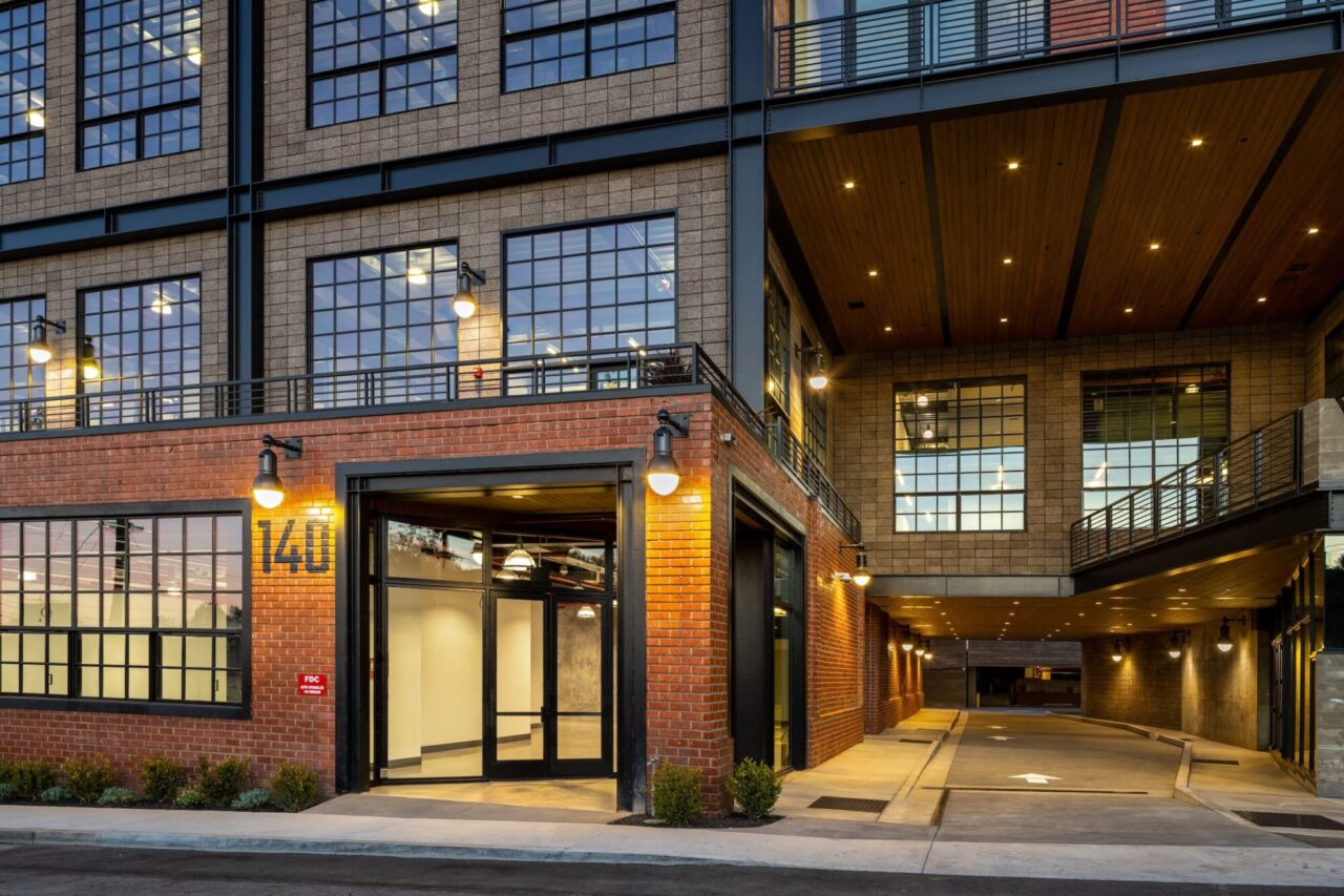 Industrial-style building numbered 140 at dusk, featuring red brick facades, gray concrete masonry structural walls, black factory-style windows, a glass entrance door, exterior sconce lighting, and an illuminated wood-ceiling parking garage entrance.
