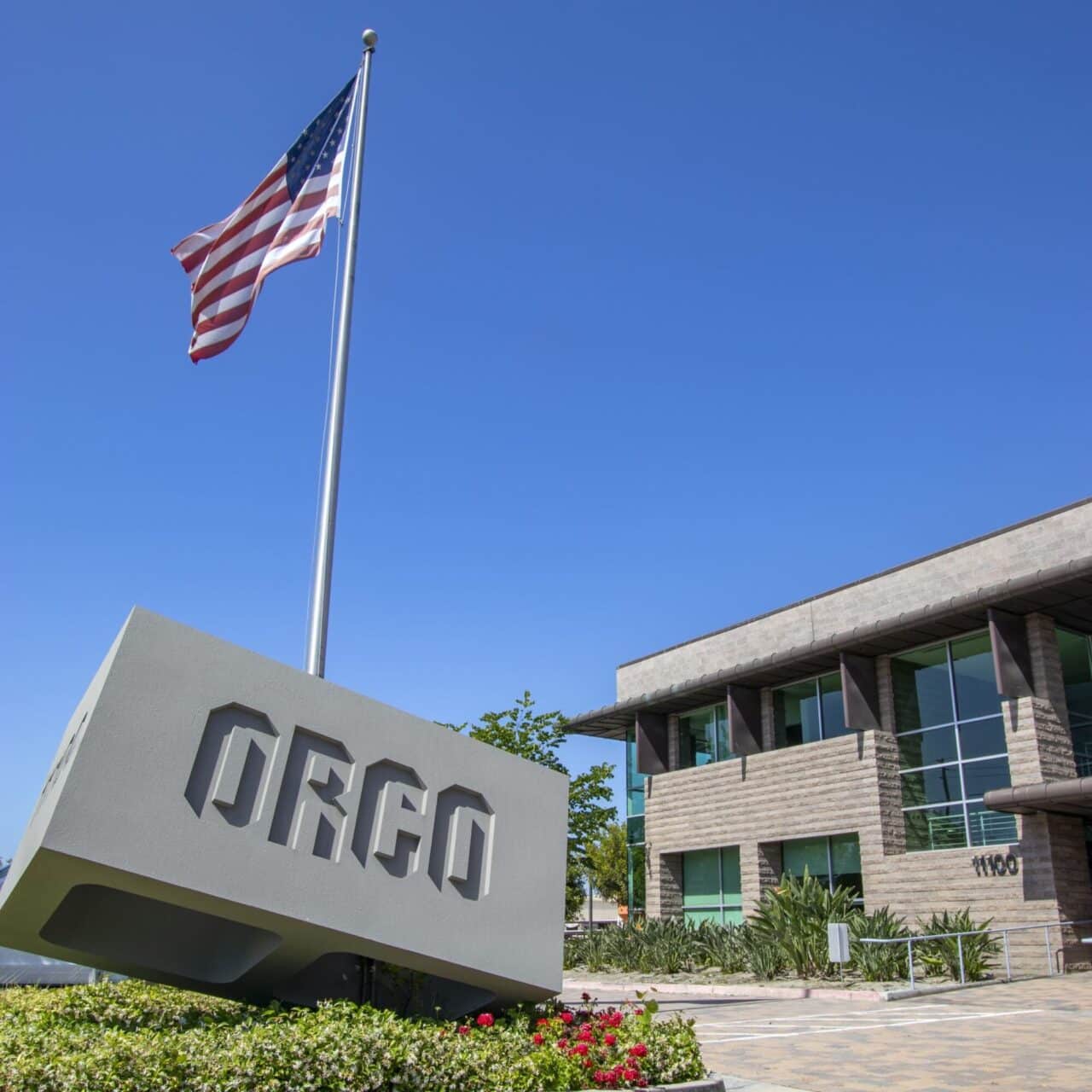 ORCO corporate headquarters entrance with a large concrete monument sign, American flag on a tall pole, and a textured stone office building numbered 11100 in the background under a clear blue sky.