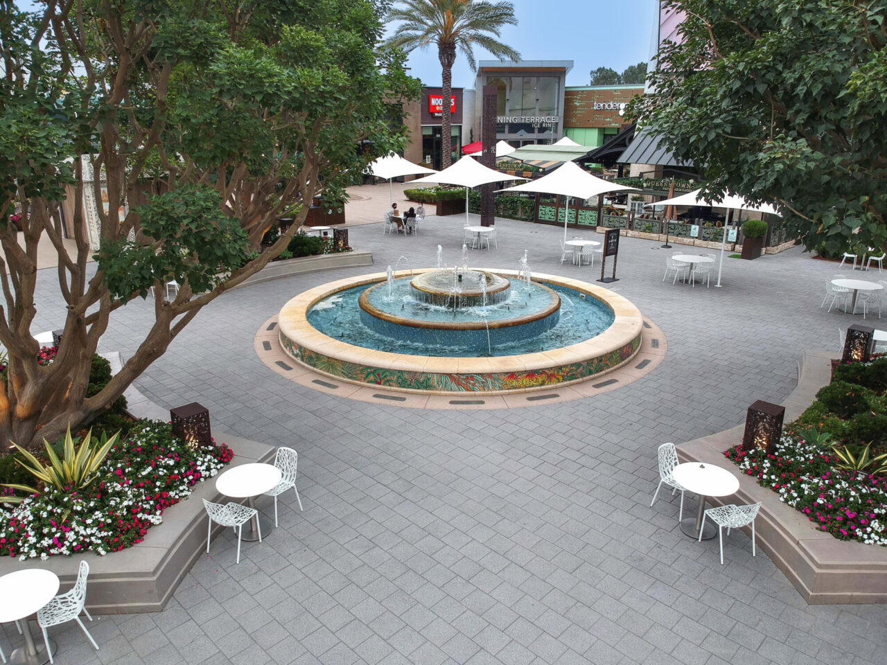 Outdoor shopping center plaza with square pavers and a tiered tile-mosaic fountain as the centerpiece, surrounded by white bistro tables and chairs, colorful flower beds, mature trees, and dining terraces with market umbrellas.