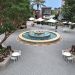Outdoor shopping center plaza with square pavers and a tiered tile-mosaic fountain as the centerpiece, surrounded by white bistro tables and chairs, colorful flower beds, mature trees, and dining terraces with market umbrellas.