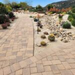 Paver walkway winding past a dry riverbed rock garden with barrel cactus and desert plants framed by hills in the background.