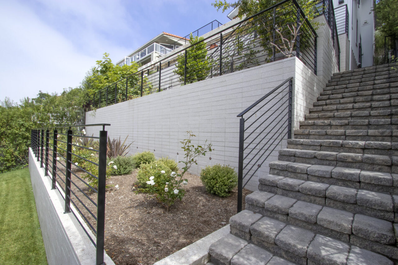 Paver stairway with black metal railings ascending a terraced hillside, bordered by a veneer tarraced wall and a modern home above.