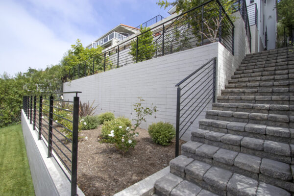 Paver stairway with black metal railings ascending a terraced hillside, bordered by a veneer tarraced wall and a modern home above.