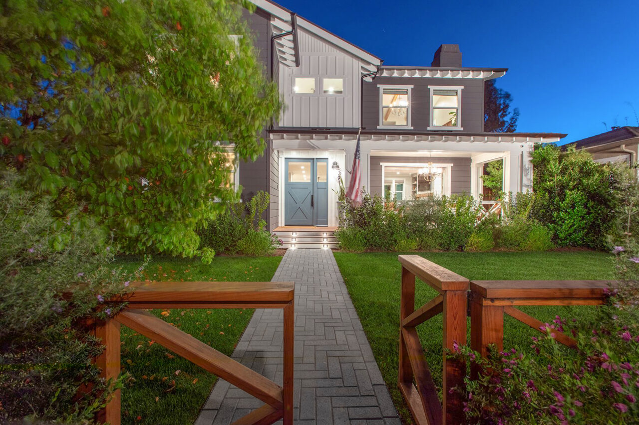 Herringbone paver walkway leading through a lush front lawn to a two-story farmhouse-style home glowing with evening light.