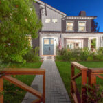 Herringbone paver walkway leading through a lush front lawn to a two-story farmhouse-style home glowing with evening light.