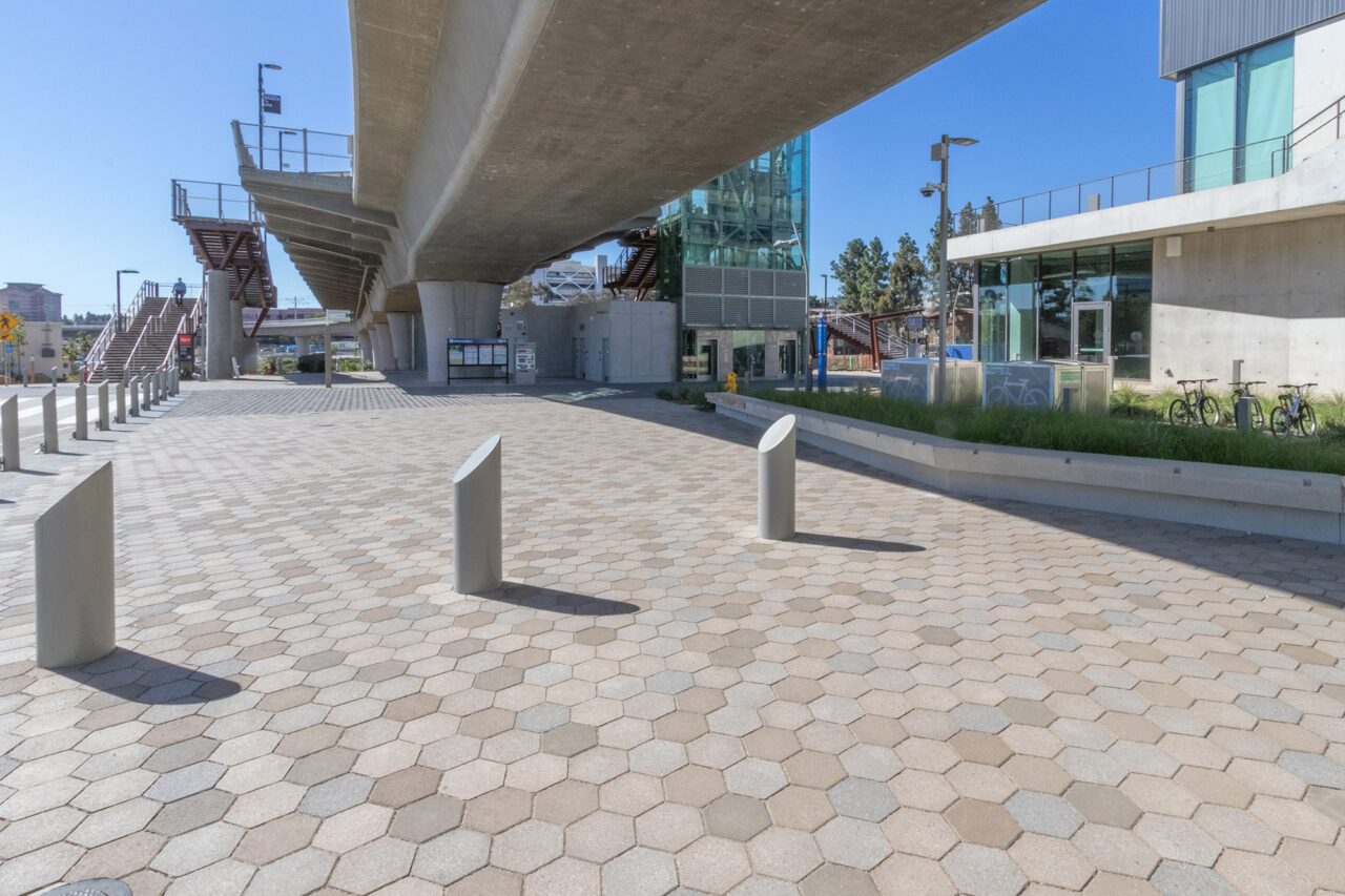 Urban plaza with hexagonal paver flooring beneath an elevated concrete transit overpass, featuring angled bollards, staircases to the platform above, bike racks, and a glass-walled transit building in the background.