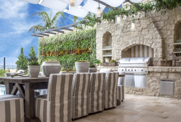 Outdoor dining area with striped slipcovered chairs beside a stone outdoor kitchen with built-in grill and a living green wall.