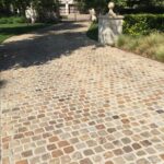 Cobblestone driveway shaded by trees, leading to an ornate iron entry gate flanked by a stone pillar with a decorative finial.