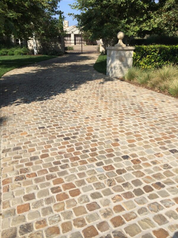 Cobblestone driveway shaded by trees, leading to an ornate iron entry gate flanked by a stone pillar with a decorative finial.
