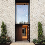 Wood-look porcelain tile entryway leading to a tall dark wood door set in a stacked-stone facade, flanked by potted conifers and flowering plants.