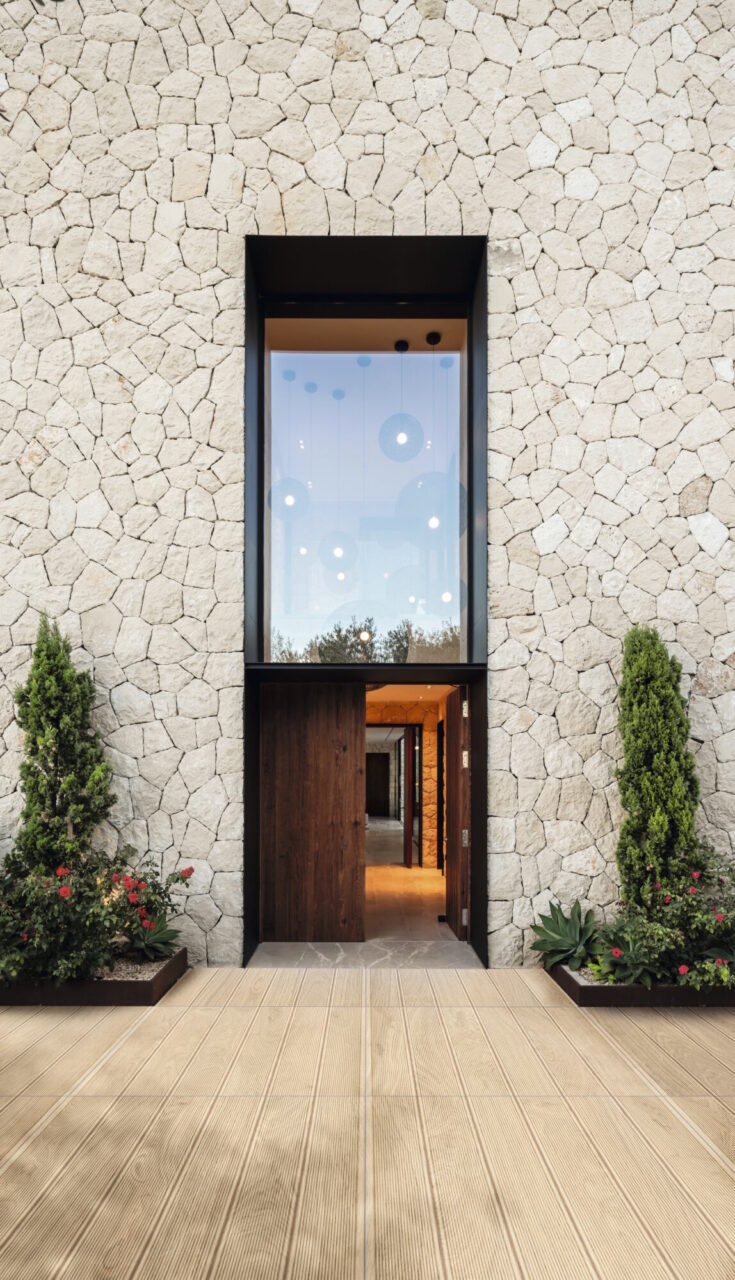Wood-look porcelain tile entryway leading to a tall dark wood door set in a stacked-stone facade, flanked by potted conifers and flowering plants.