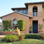 Two-story brick and stucco Mediterranean-style home with tile roof, blue shutters, and a dark front door, featuring a landscaped entrance with brick steps, potted agave plants with American flags, manicured hedges, and an attached garage.