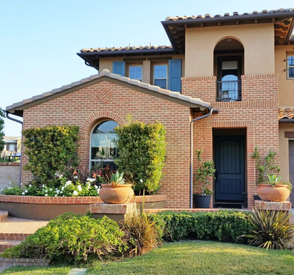 Two-story brick and stucco Mediterranean-style home with tile roof, blue shutters, and a dark front door, featuring a landscaped entrance with brick steps, potted agave plants with American flags, manicured hedges, and an attached garage.