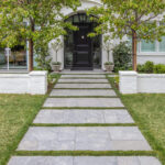 Natural stone paver walkway with grass joints leading to a black front door on a white colonial-style home, framed by trees and low brick walls.