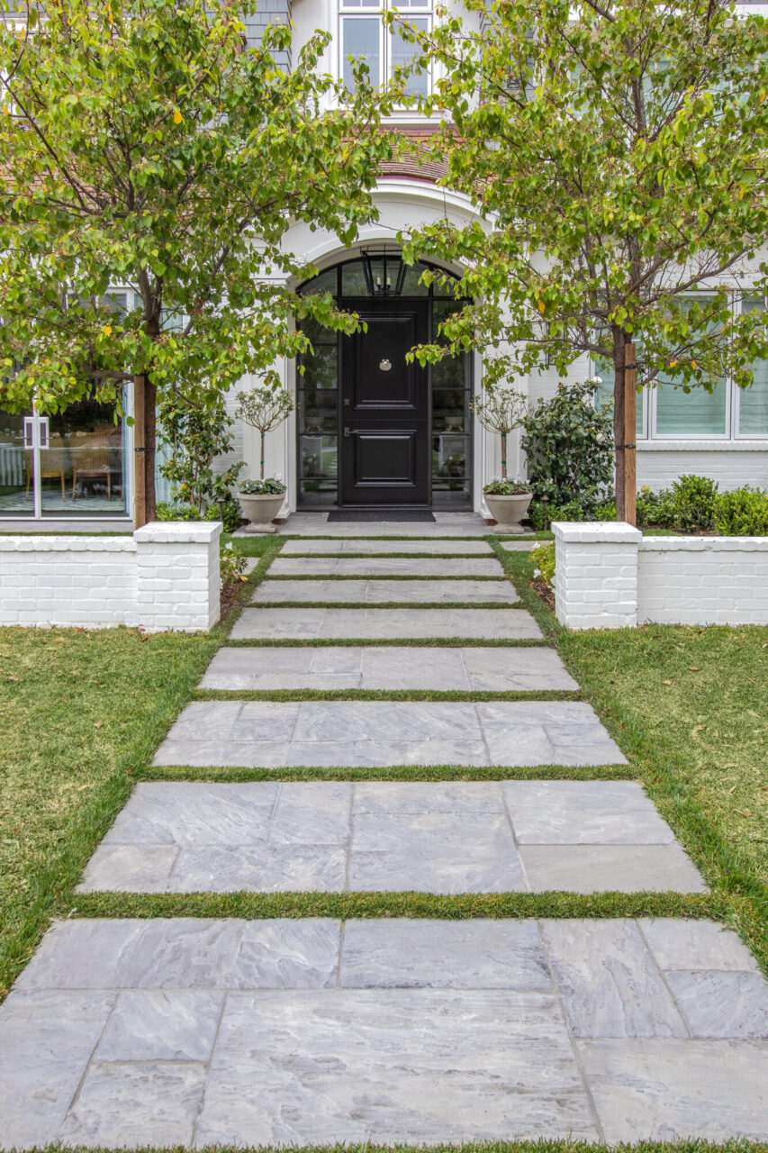 Natural stone paver walkway with grass joints leading to a black front door on a white colonial-style home, framed by trees and low brick walls.