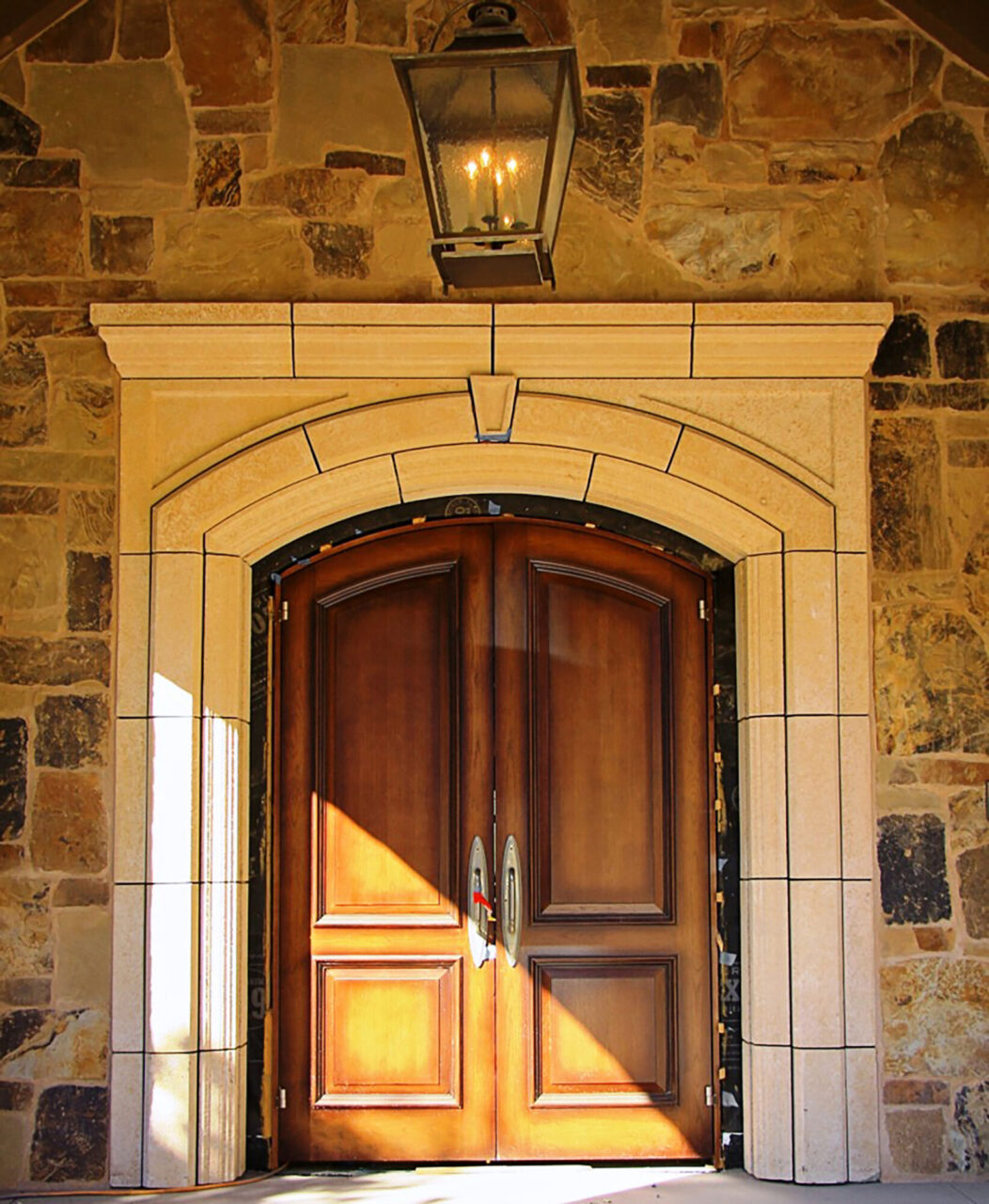 Grand double front door in rich stained wood with an arched limestone surround, curved metal handles, and a hanging lantern-style light fixture, set into a stacked stone facade.